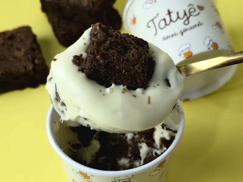 Pote da Tatyê com creme e um pedaço de brownie por cima, com colher dourada e brownies ao lado