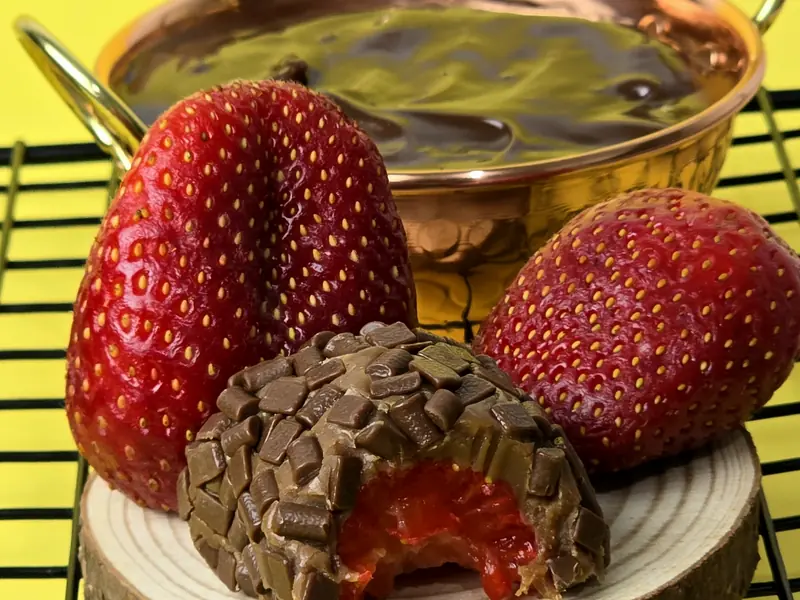 Brigadeiro de morango mordido, com o recheio à vista, sobre disco de madeira, ao lado de morangos inteiros