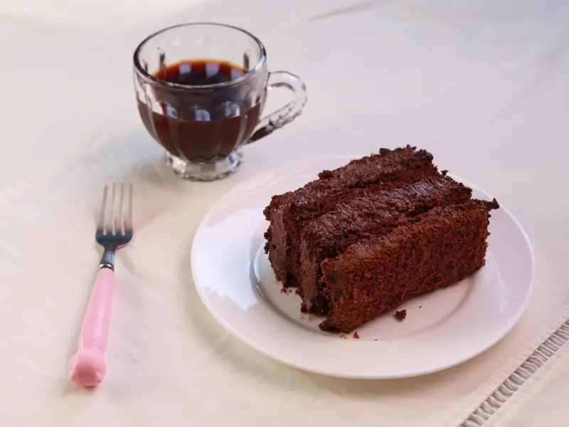 Fatia de bolo de chocolate em camadas num prato branco, com garfo de cabo rosa e xícara de vidro ao fundo